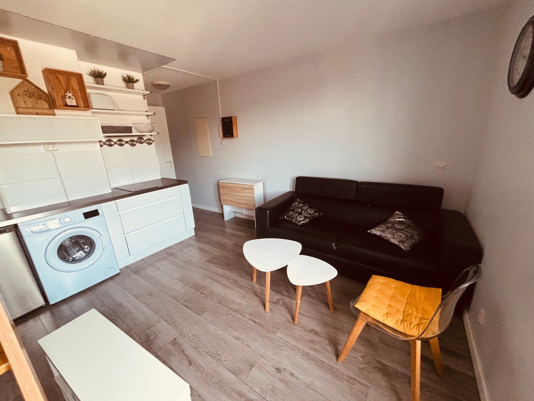 A compact living space with a kitchen, a black sofa, two white side tables, and a wooden stool.