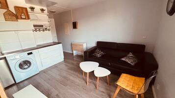 A compact living space with a kitchen, a black sofa, two white side tables, and a wooden stool.