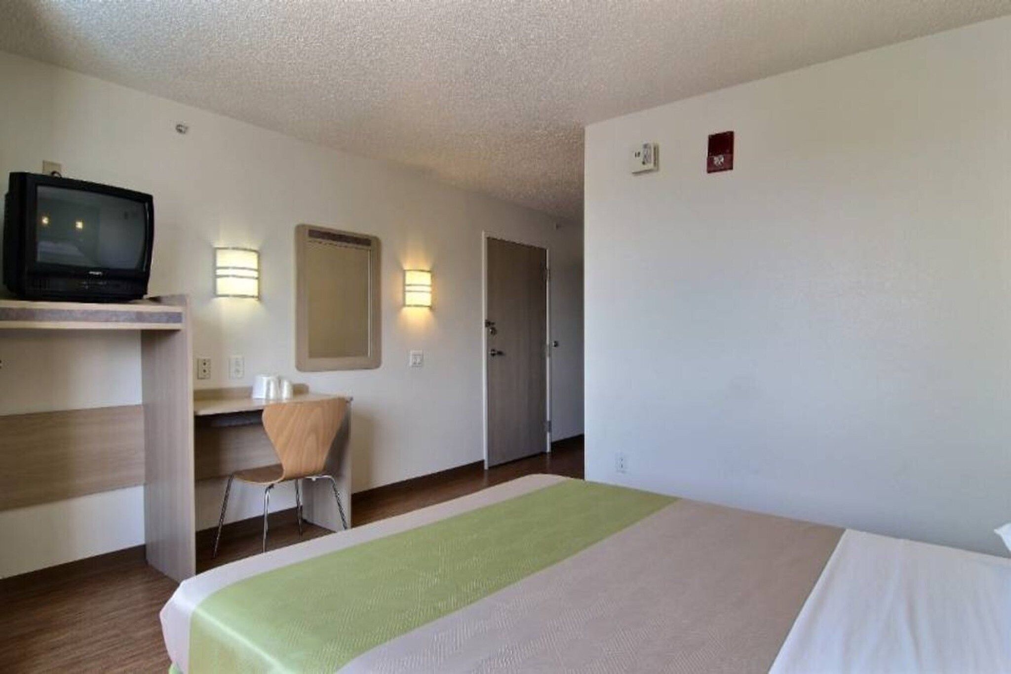 A hotel room with a bed, a desk with a chair, a television, and two wall-mounted light fixtures.