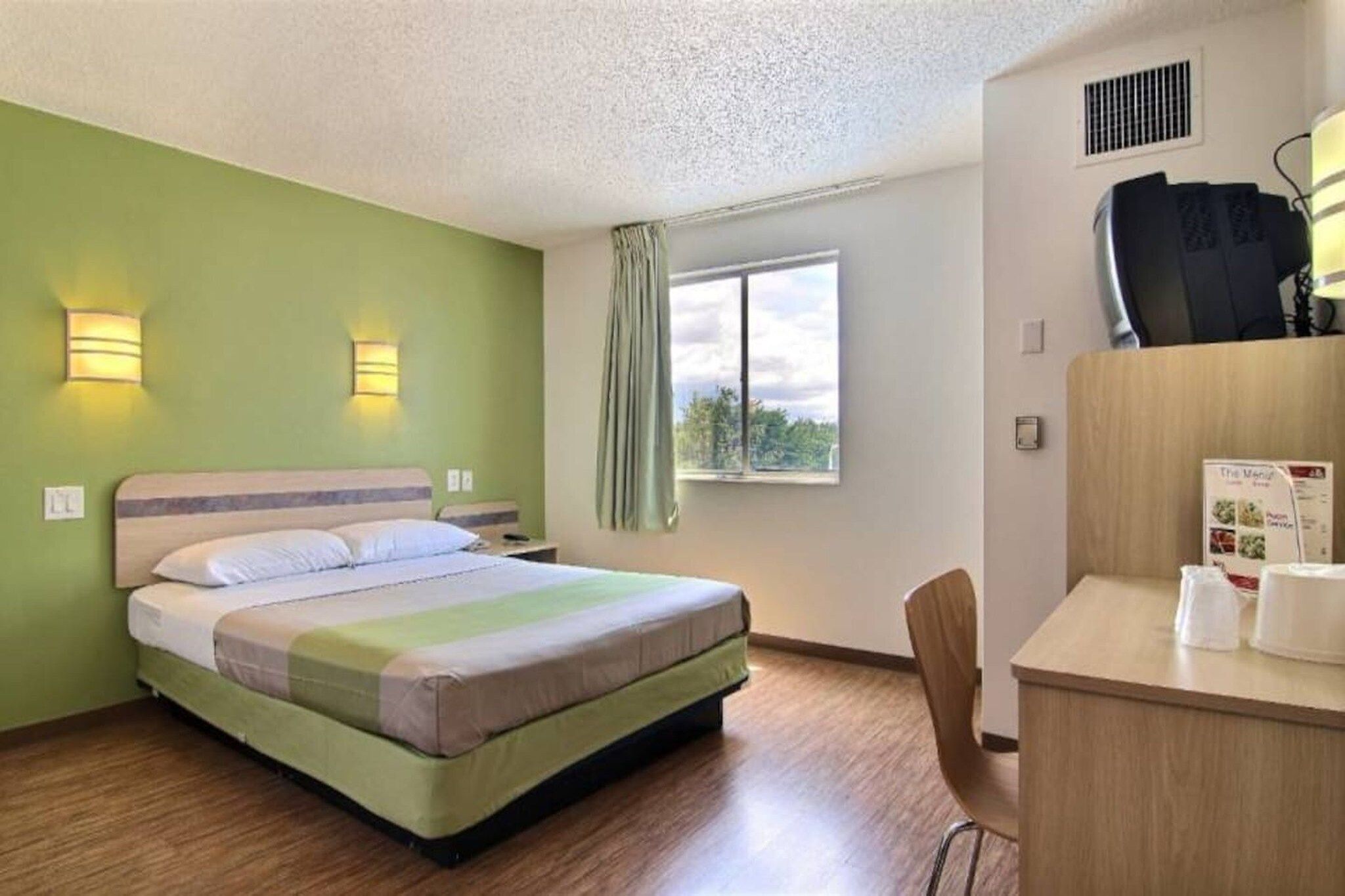 A hotel room with a bed, a desk with a chair, a window with curtains, and a wall-mounted air conditioner.