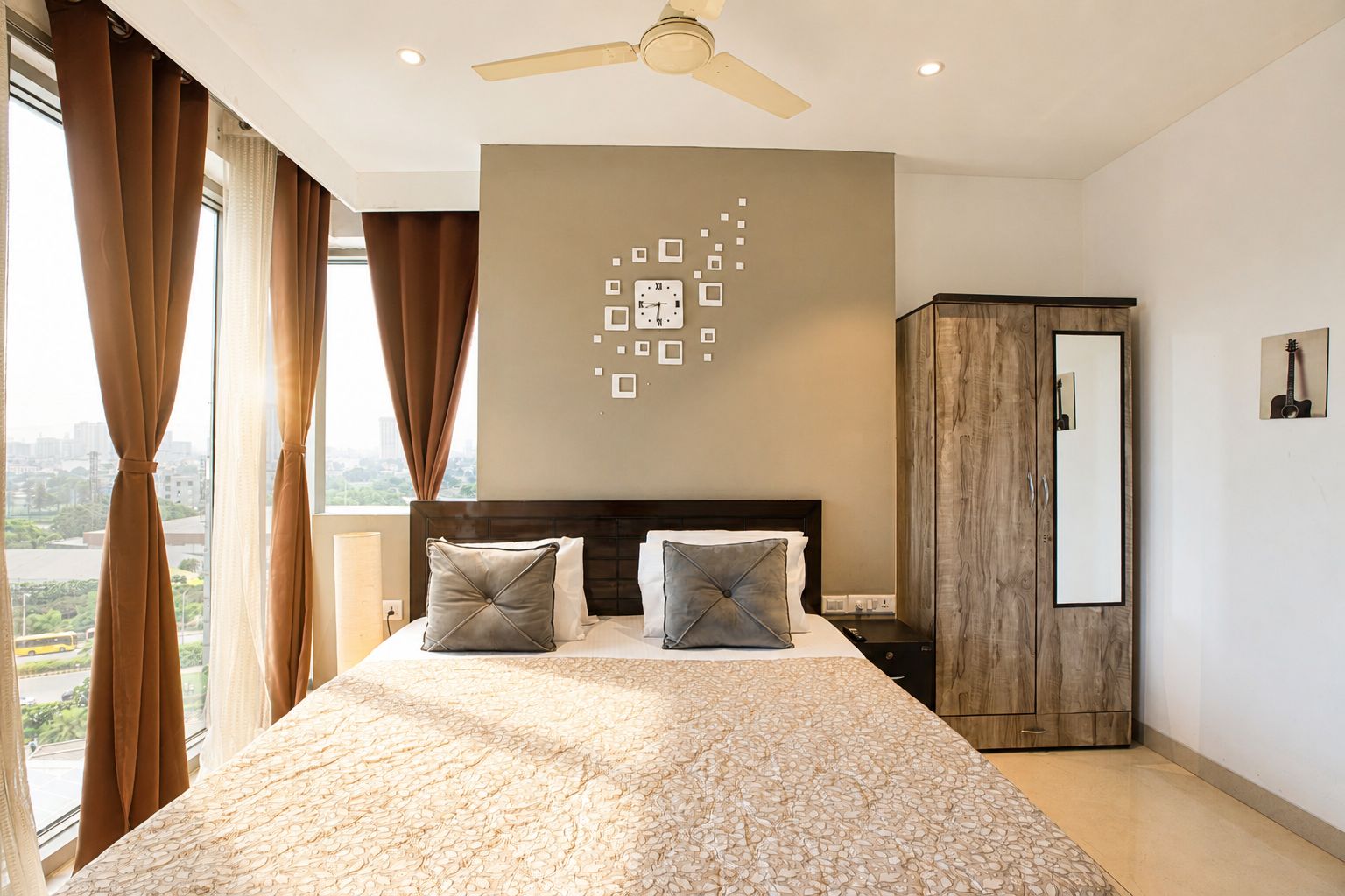A hotel room with a bed, curtains, a ceiling fan, a wall clock, a wardrobe, and a city view.