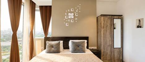 A hotel room with a bed, curtains, a ceiling fan, a wall clock, a wardrobe, and a city view.