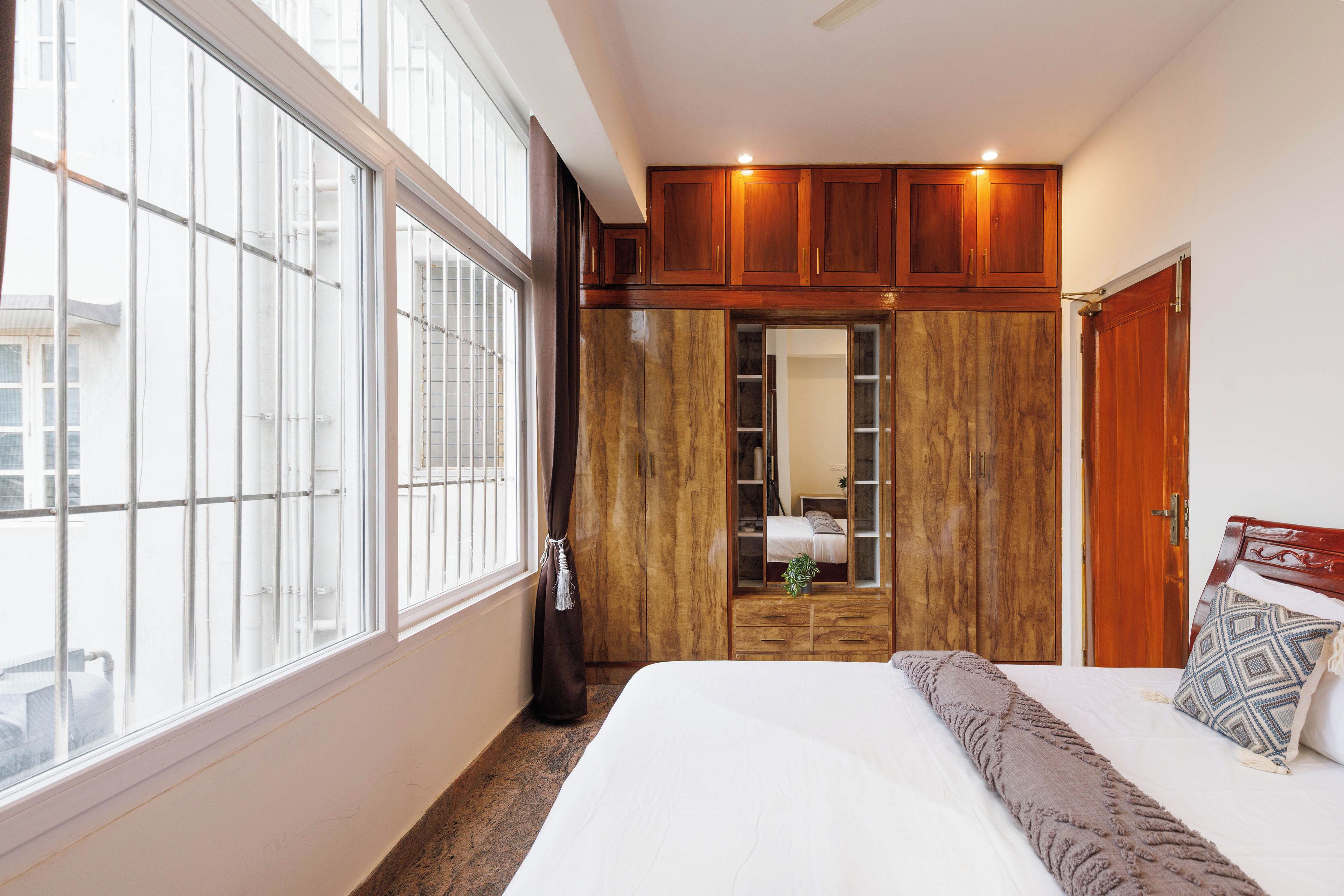 A bedroom with a large window, wooden wardrobe, and a bed with a patterned pillow.