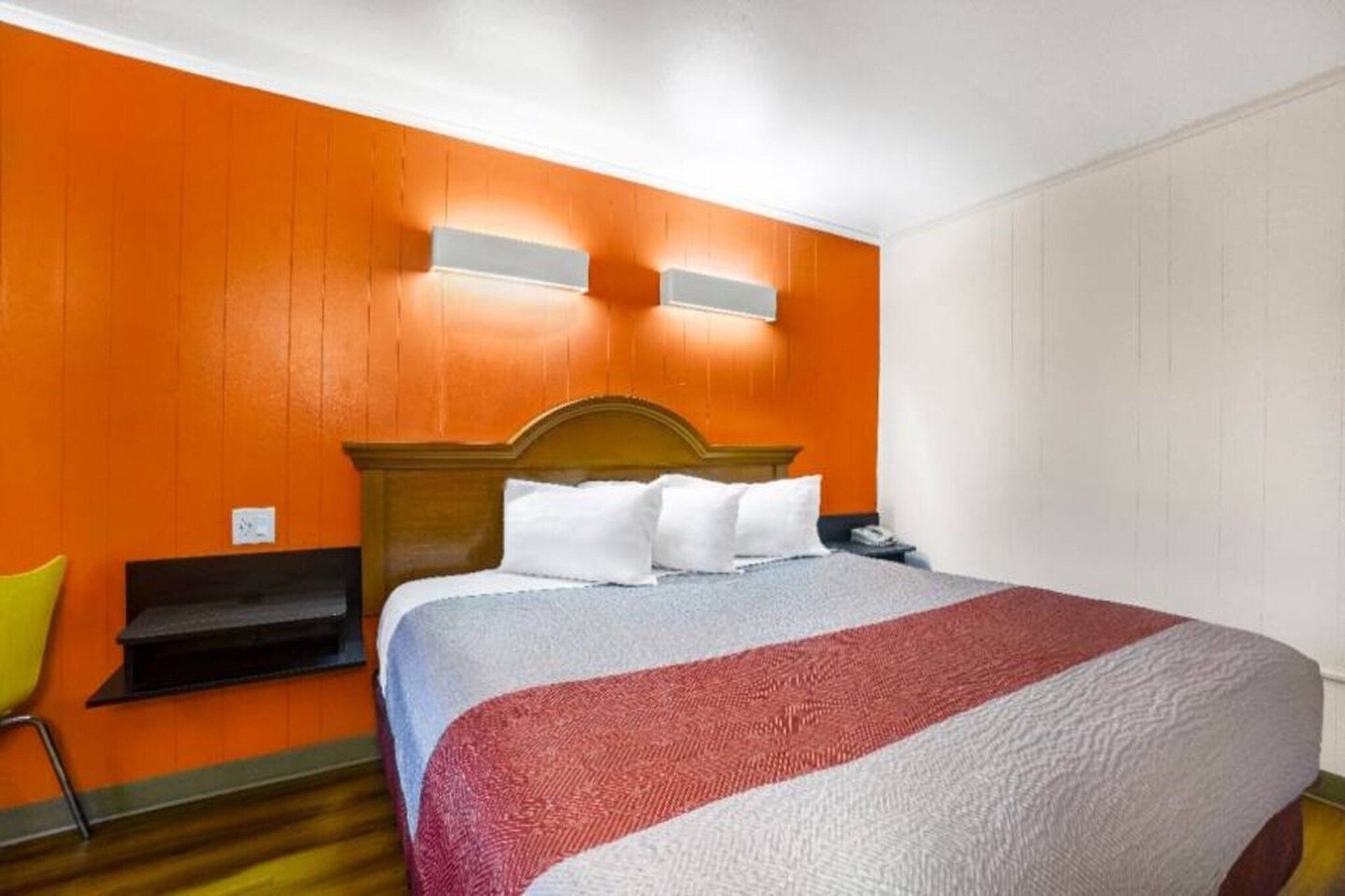 Une chambre d’hôtel avec une tête de lit en bois, un lit avec une literie blanche et rouge, une chaise jaune et un mur orange.