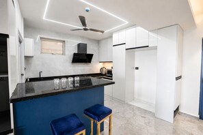 A modern kitchen with a black countertop, white cabinets, a ceiling fan, and a blue bar stool. - The Canvas by Redolive 2bhkgachibowli Hitech (Hyderabad)