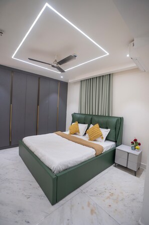 Une chambre moderne avec un lit vert, du linge de lit blanc, un ventilateur de plafond et une table de chevet ornée d’un vase de fleurs.