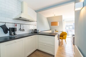 A modern kitchen with white cabinets, a black countertop, and a built-in stove. There is a yellow chair in the dining area and a large window providing natural light. - Casa Karline - Casa Karline – Relaxed Living with a View of Sylt’s Greenery (Sylt)