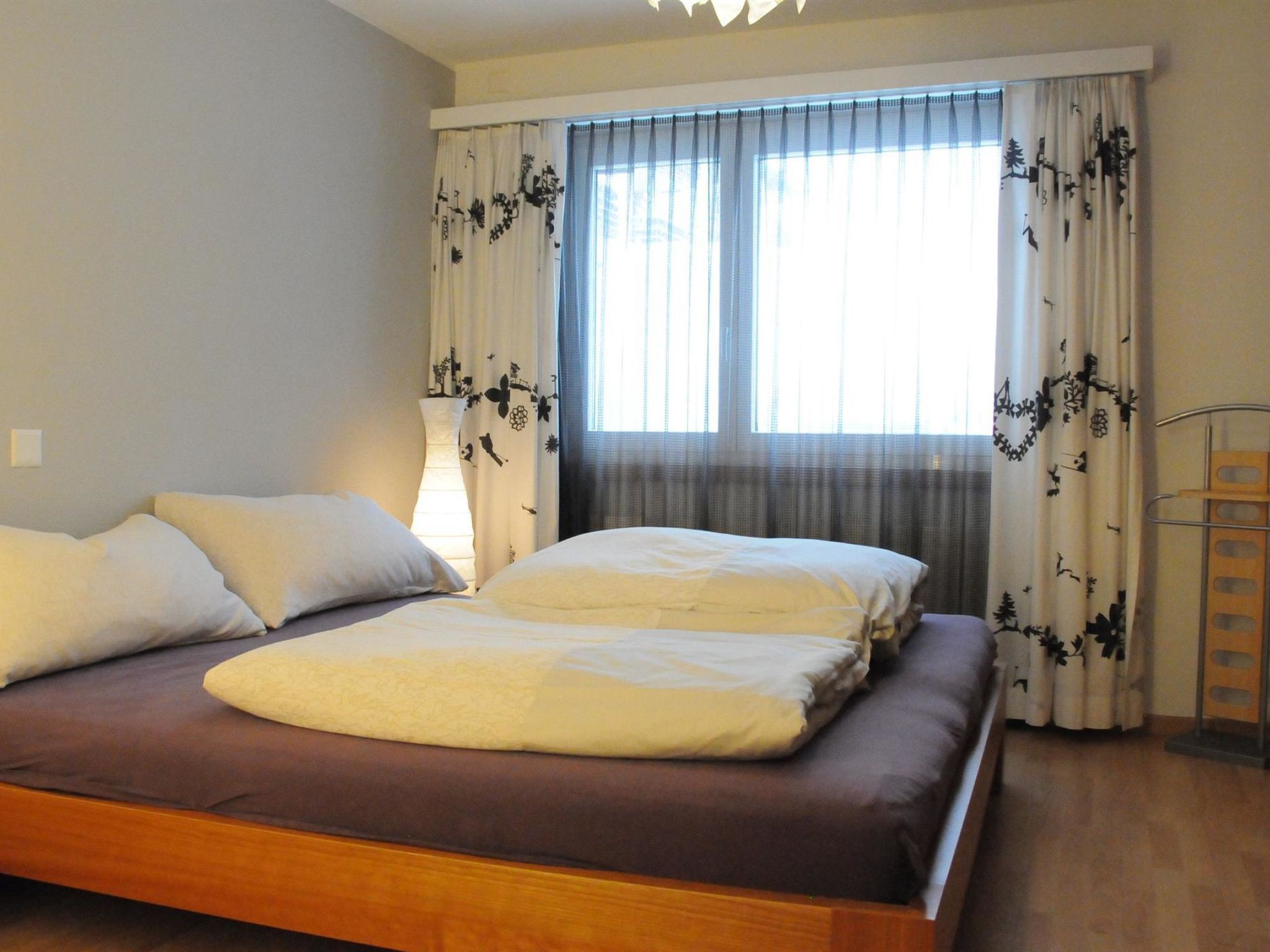 Een slaapkamer met een bed, kussens, een nachtkastje, een lamp en gordijnen met een bloemenprint.