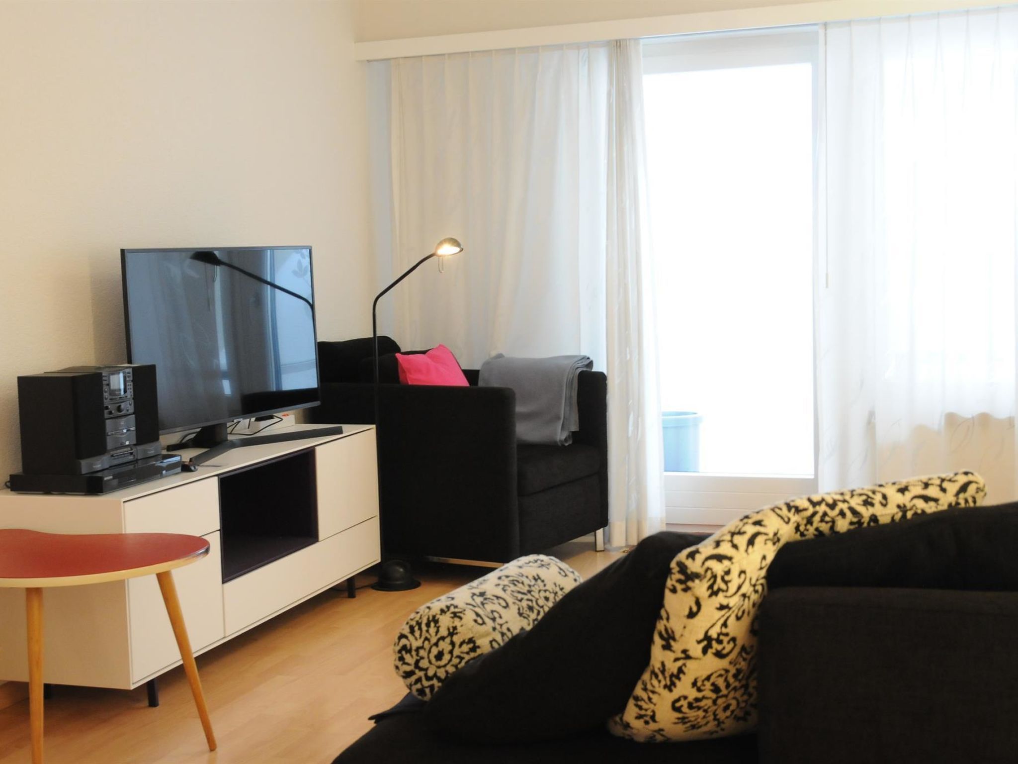 A modern living room with a flat-screen TV, a black sofa, a red chair, and a small table.