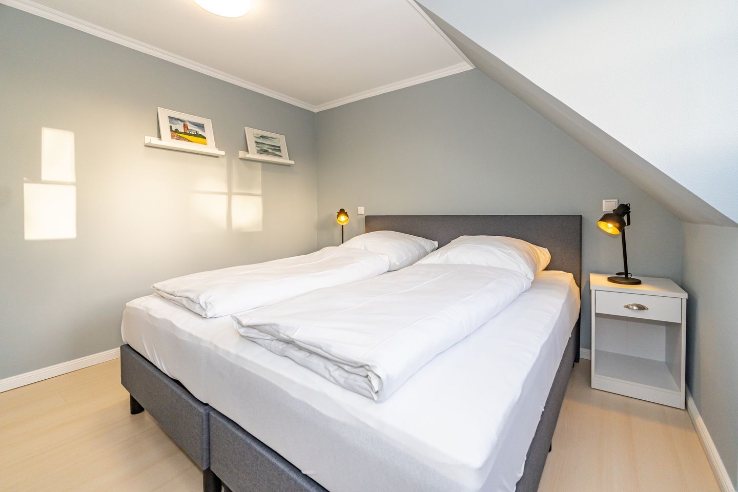 Een netjes opgemaakt tweepersoonsbed met witte lakens, een nachtkastje met een lamp, en twee ingelijste foto's aan de muur.