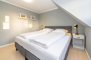 Ein ordentlich bezogenes Doppelbett mit weißen Bettwäsche, ein Nachttisch mit Lampe und zwei gerahmte Bilder an der Wand.
