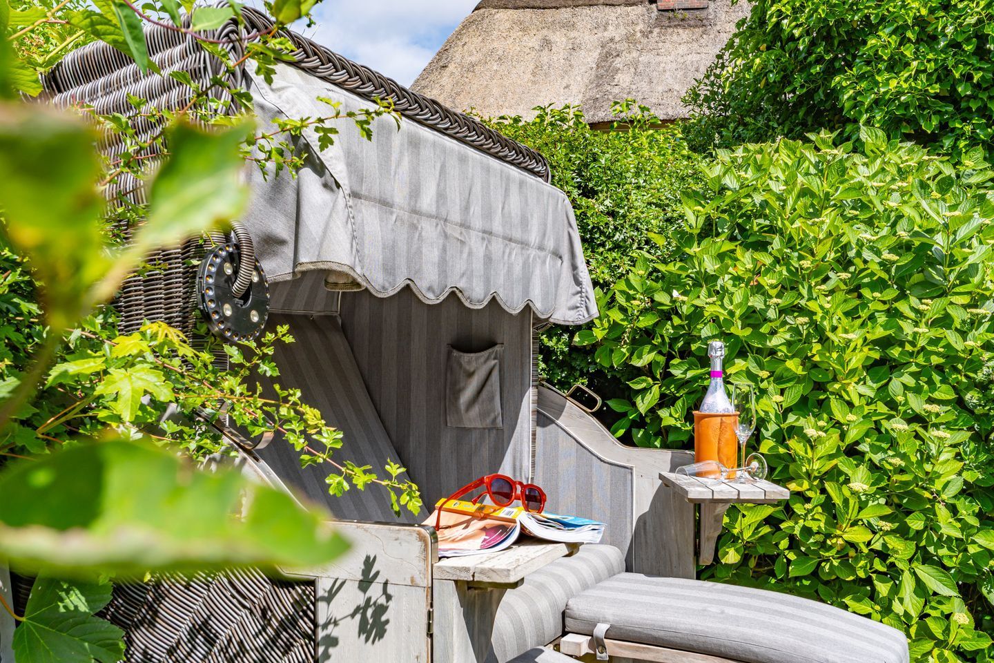 A cozy outdoor seating area with a table, a bottle, and glasses, surrounded by lush greenery and a traditional thatched roof structure.
