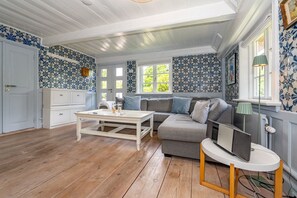 A room with a wooden floor, a white table, a grey sofa, and a window with a view of greenery. - Kapitänshuis - Kapitänshuis – a Frisian island home full of character (Sylt)