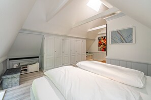 Ein Schlafzimmer mit schrägem Dach, einem Schlafplatz in der Mansarde, einem Schreibtisch und einem großen Fenster.