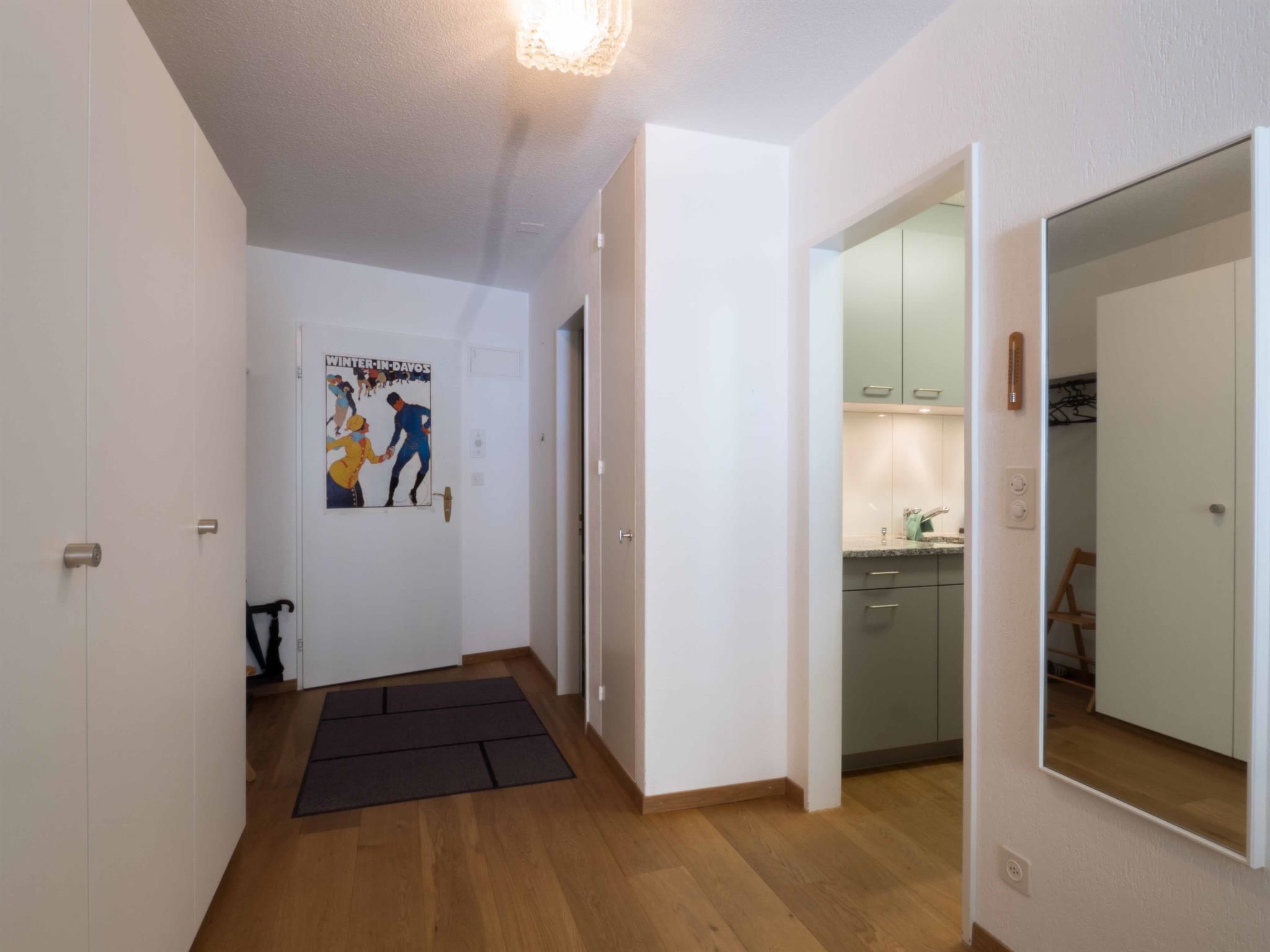Un couloir avec un plancher en bois, une affiche accrochée au mur et un coin cuisine visible à travers une porte ouverte.