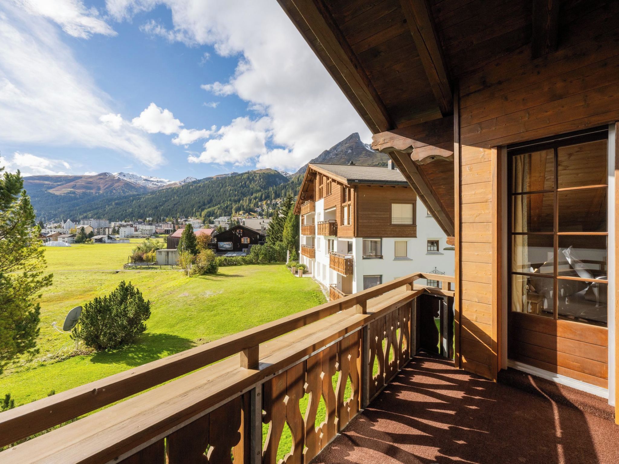 A wooden balcony with a view of a mountain, a grassy field, and a town in the distance.