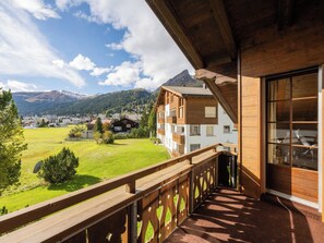 Un balcón de madera con vistas a una montaña, un campo cubierto de hierba y un pueblo a lo lejos.