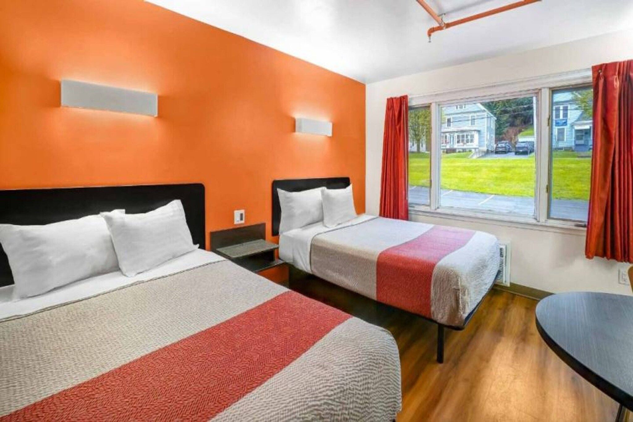A hotel room with two beds, a window with a view of a house, and a small table.