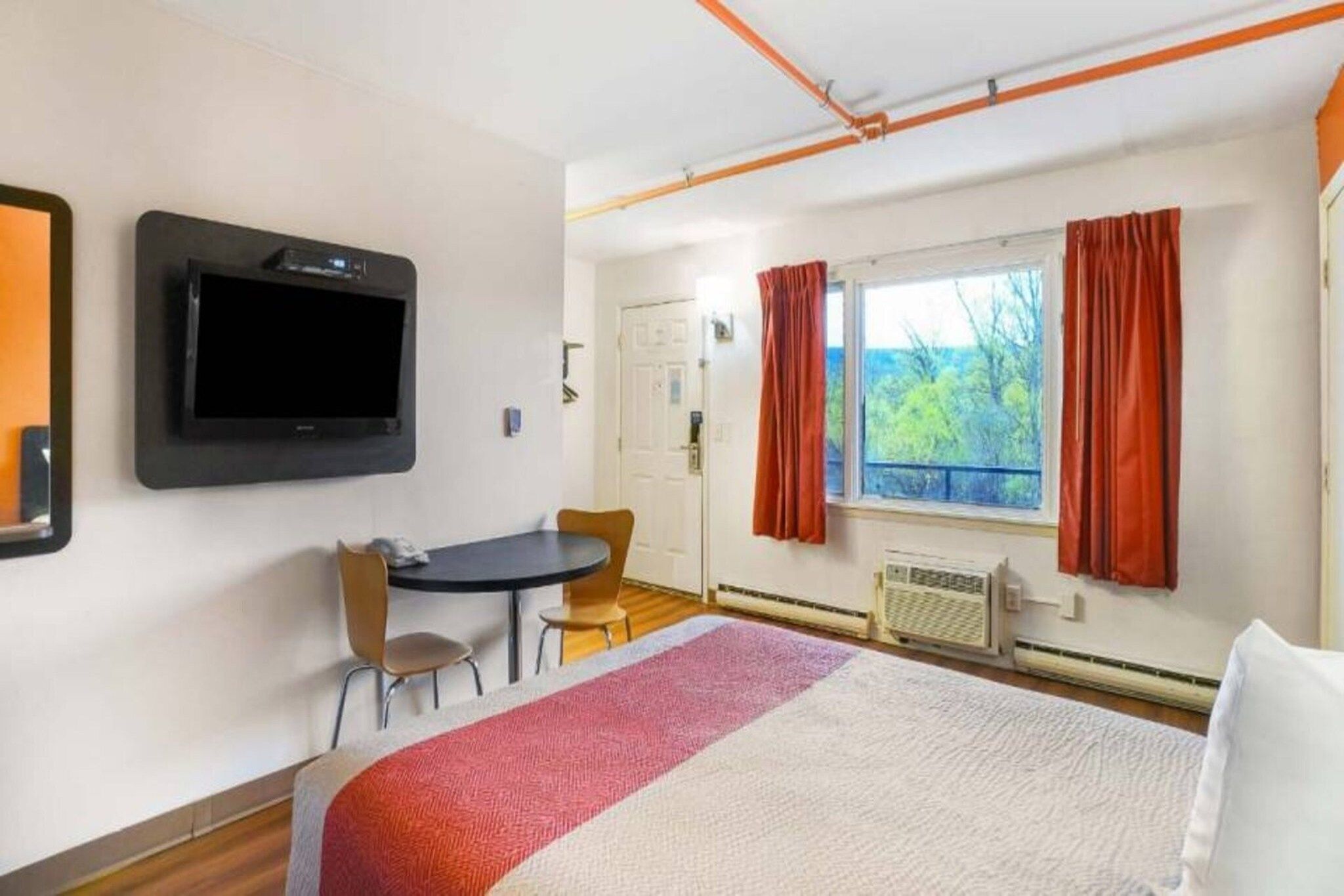 Ein Hotelzimmer mit einem Bett, einem an der Wand befestigten Fernseher, einem Esstisch mit Stühlen, einem Fenster mit Vorhängen und einem Heizkörper.