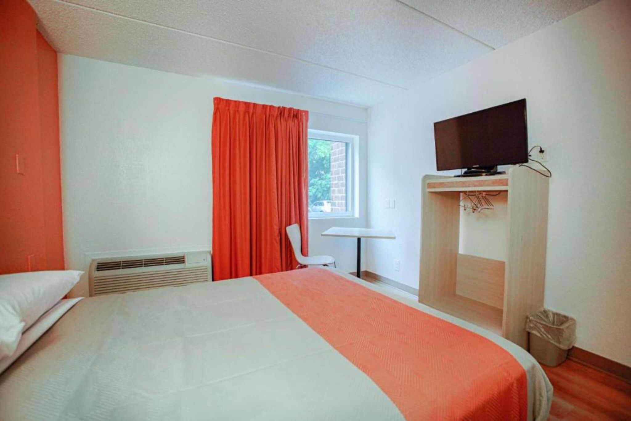 A hotel room with a bed, a television on a wooden stand, a window with curtains, and a small table.