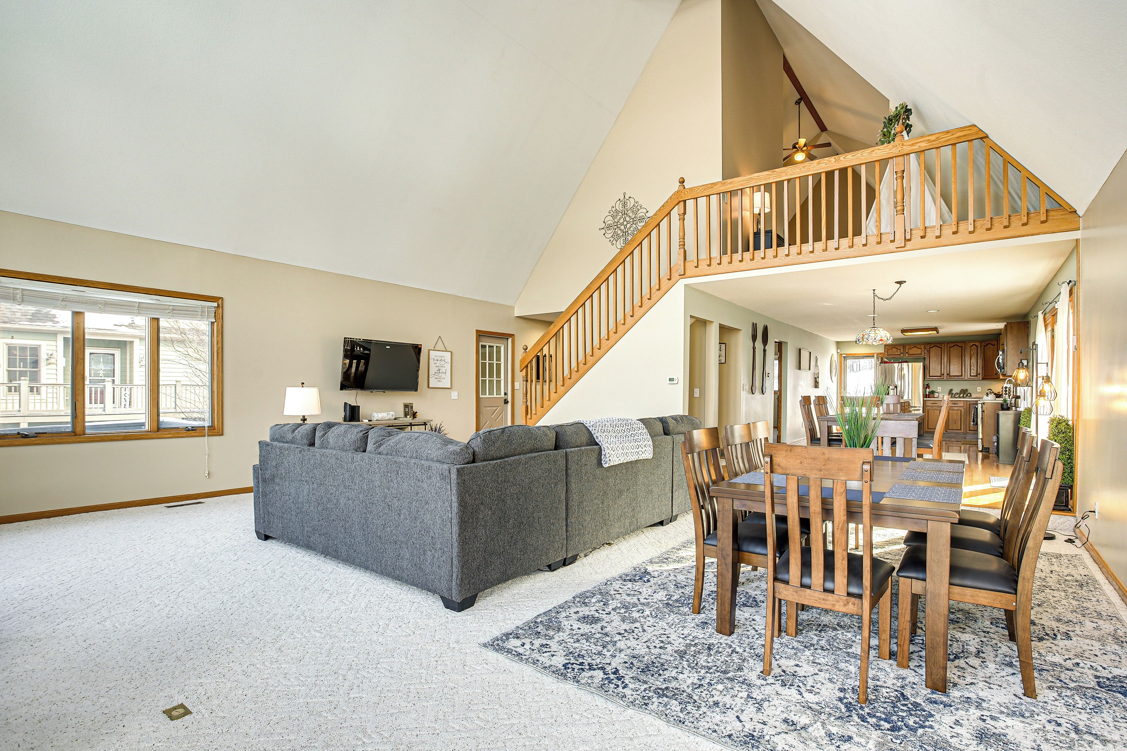 A spacious living room with a grey sectional sofa, wooden dining table with chairs, a staircase, and a kitchen in the background.
