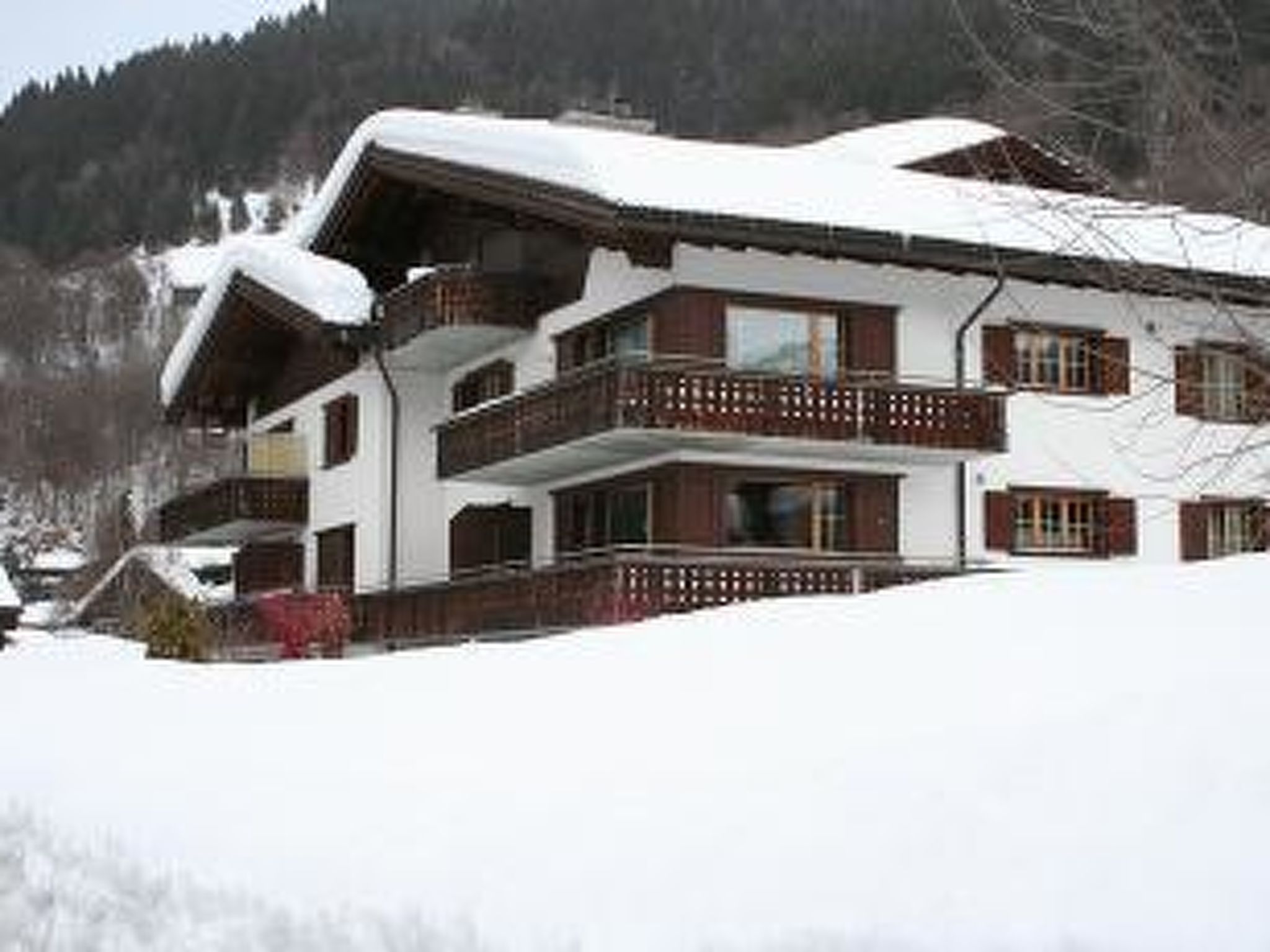 Un chalet à plusieurs étages, avec un toit enneigé, des balcons en bois et un paysage montagneux en arrière-plan.