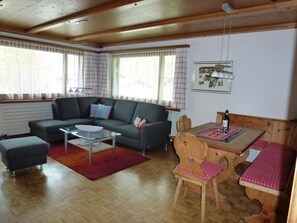 Ein gemütliches Wohnzimmer mit einer Couch, einem Glastisch, einem Holzes Esstisch mit Stühlen und einem Fenster mit Vorhängen.