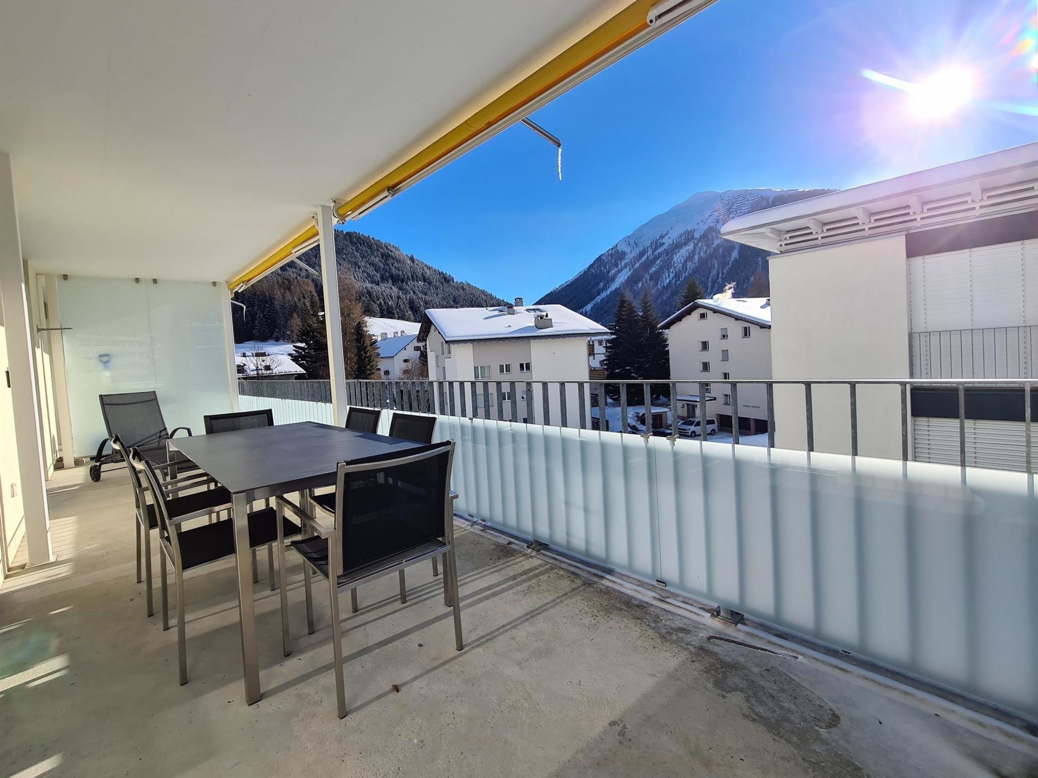 Un balcón con mesa y sillas, con vista a un paisaje montañoso y edificios.