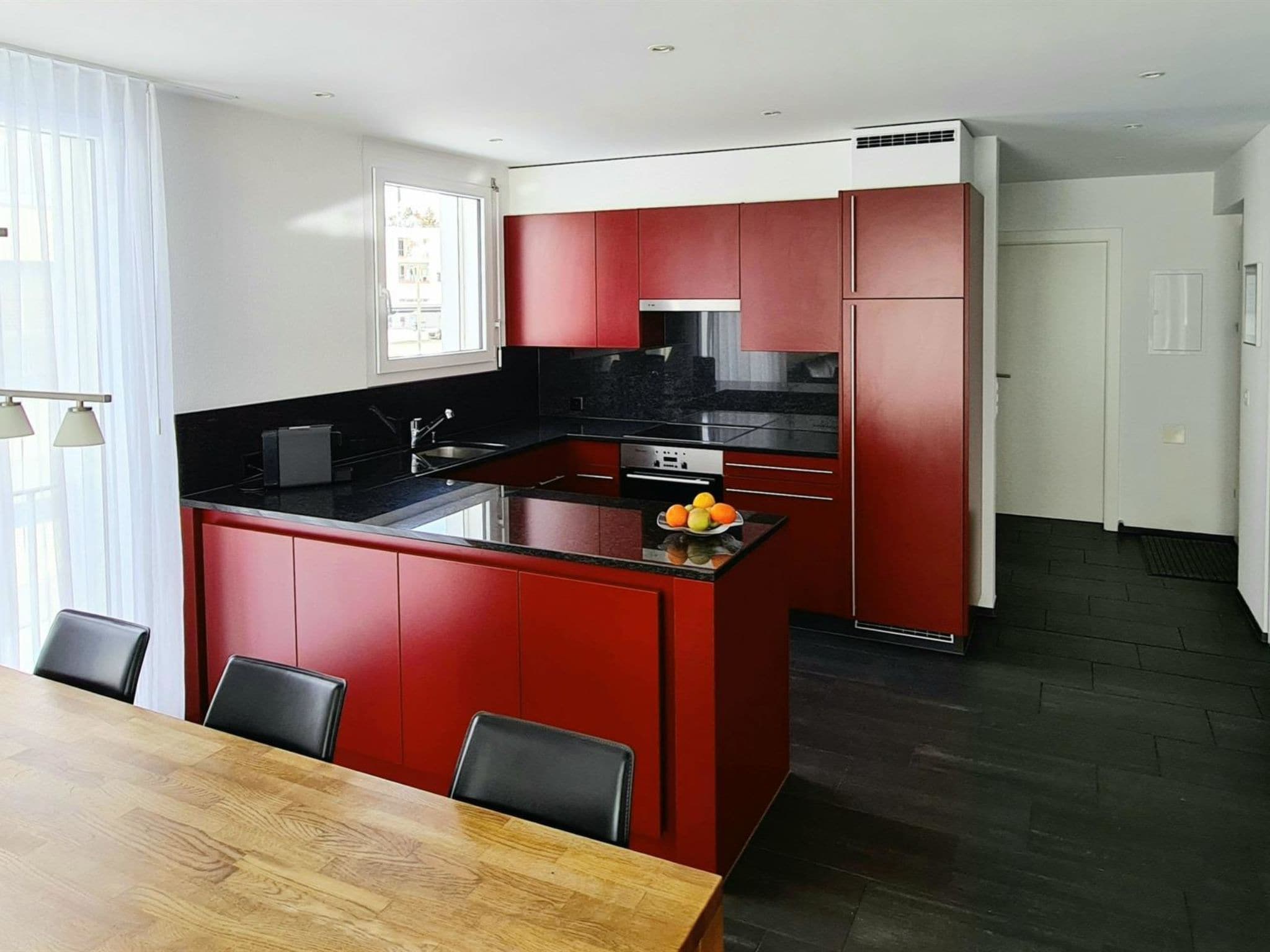 A modern kitchen with red cabinetry, a black countertop, and a central island with fruit.