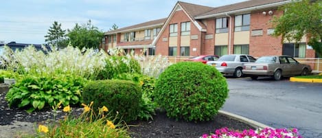 Un hotel con fachada de ladrillo, estacionamiento y un jardín ajardinado con diversas plantas y flores.