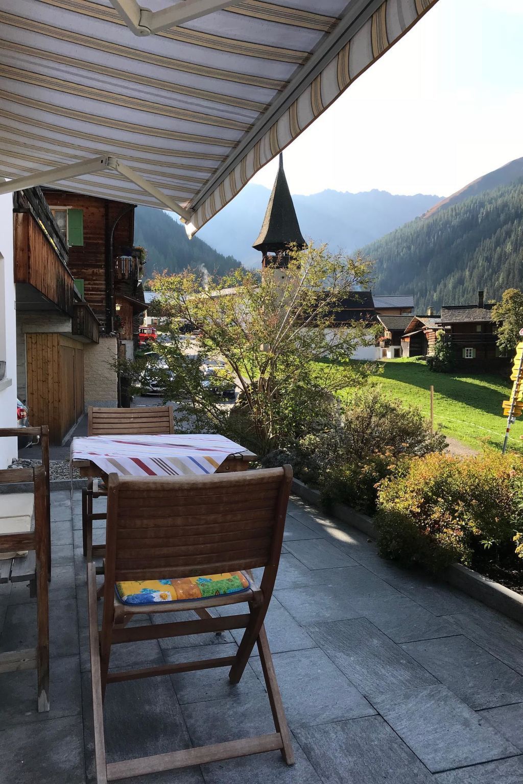 An outdoor patio with a table and chairs, overlooking a mountainous landscape with a church steeple.