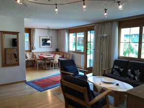 Ein geräumiges Wohnzimmer mit einem Esstisch, Sofas und einem großen Fenster mit Blick ins Freie.