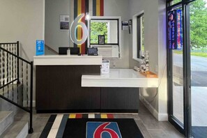 A reception desk with a digital display, a framed picture, and a window with a view of greenery. - 2 Full Beds | Non-Smoking (East Windsor)