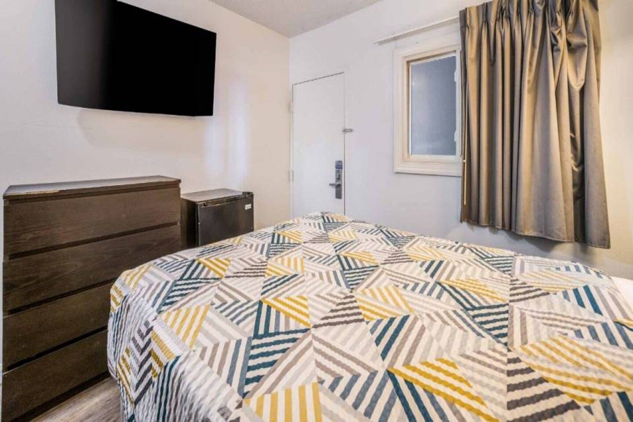 A bedroom with a bed, a dresser, a television mounted on the wall, a door, and a window with curtains.