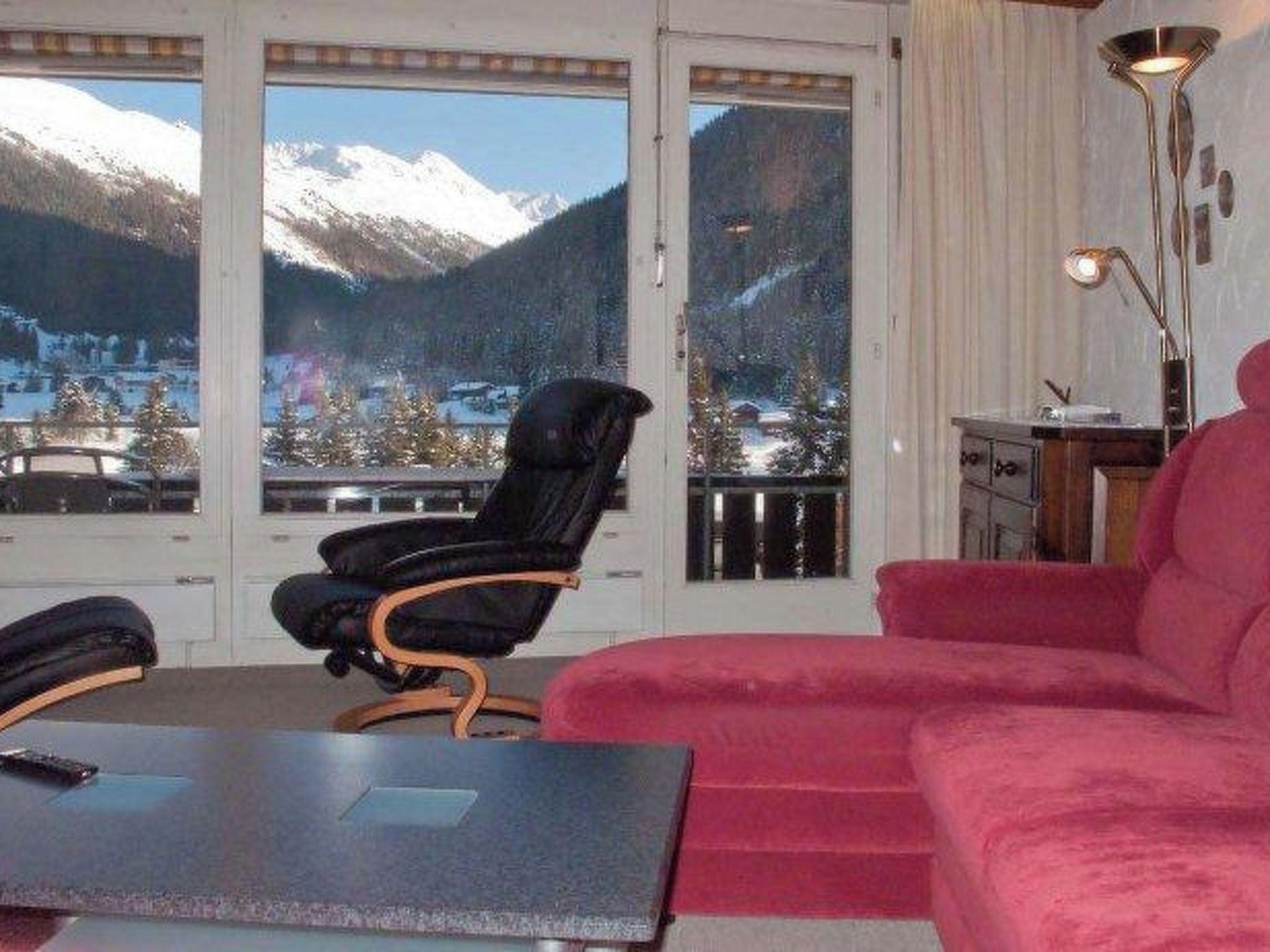 Ein Zimmer mit Blick auf schneebedeckte Berge, ein schwarzer Sessel, ein rotes Sofa und ein Schreibtisch mit Lampe.