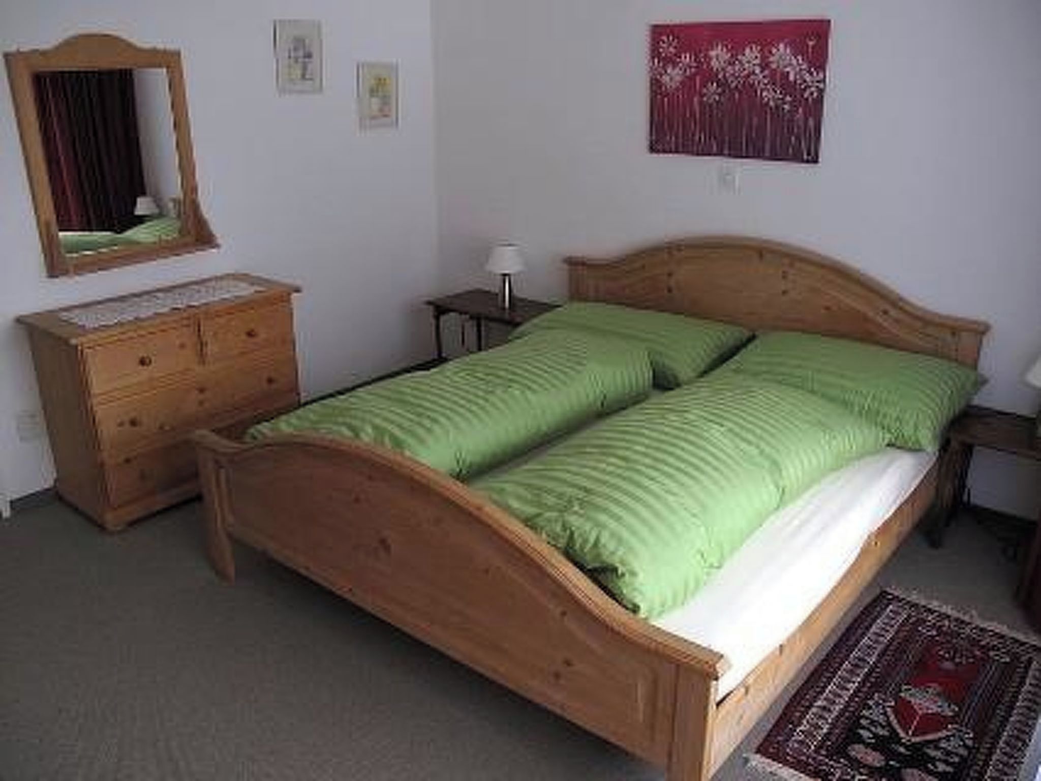 A bedroom with a wooden bed, a dresser, a nightstand, a lamp, and a wall-mounted mirror.