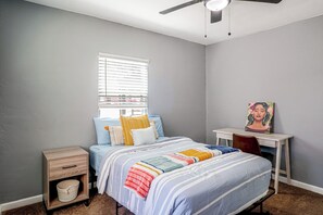 Ein Schlafzimmer mit Bett, Schreibtisch, Stuhl, einem Fenster mit Jalousien und einem Deckenventilator.