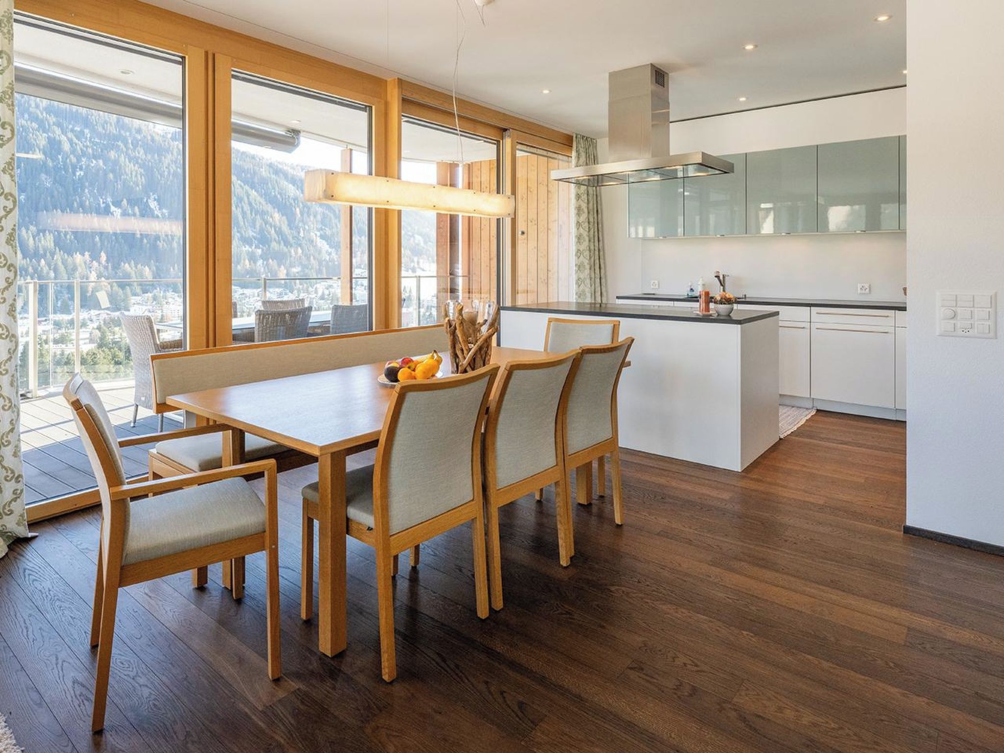 Una cocina moderna con una isla central, mesa de comedor de madera con sillas y grandes ventanales con vistas a las montañas.