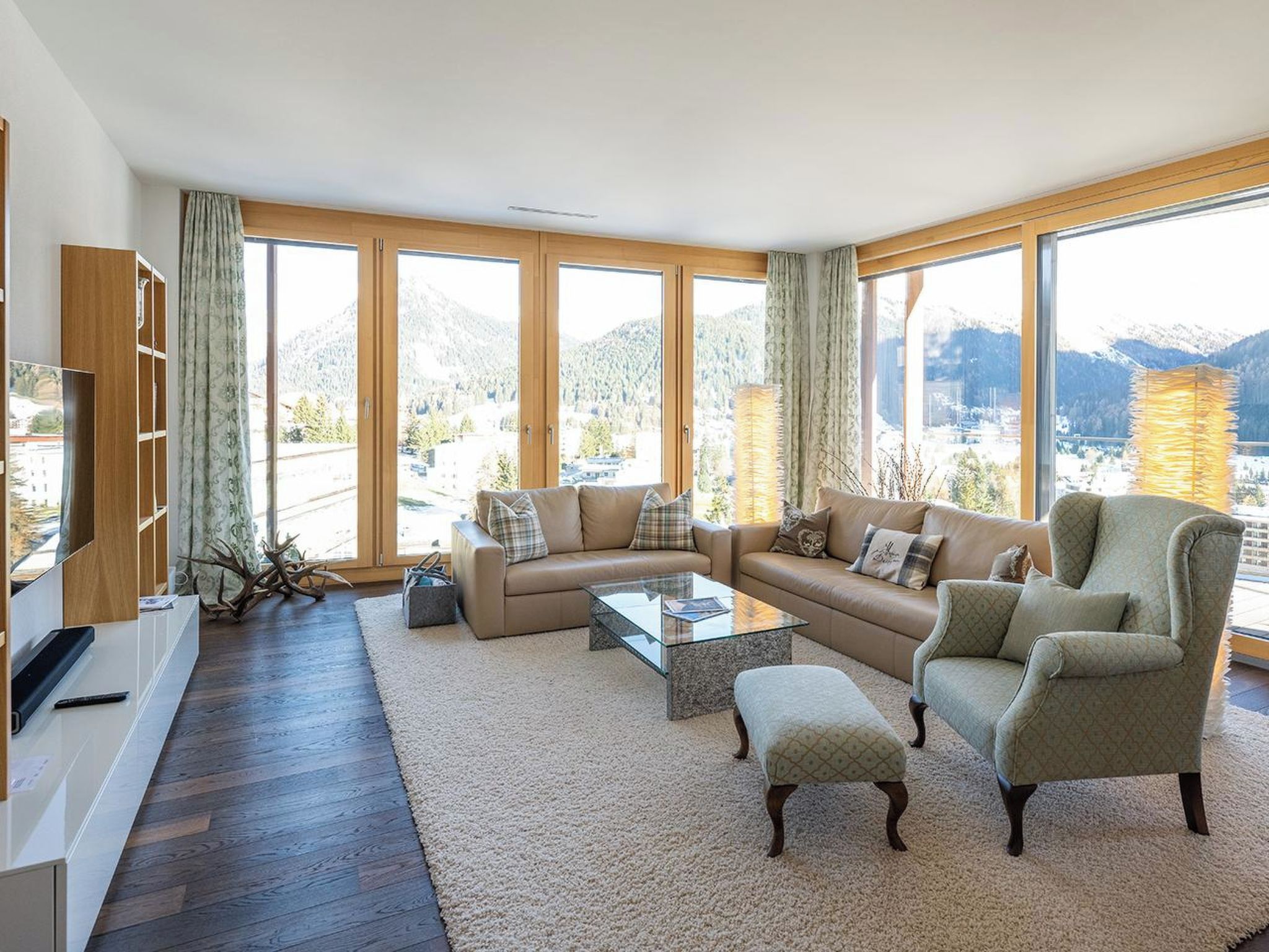 A spacious living room with a sofa, armchair, and coffee table, large windows with a mountain view, and a wooden TV stand.