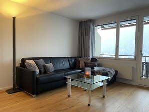 A modern living room with a black leather sofa, a glass coffee table, and a view through a large window. - Rex 33 Appartement/Fewo, Dusche oder Bad, WC 2½-Zimmerwohnung by Interhome (Davos)