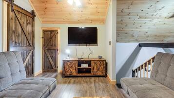 A cozy living room with a wooden ceiling, a large TV mounted on the wall, and a rustic wooden cabinet.