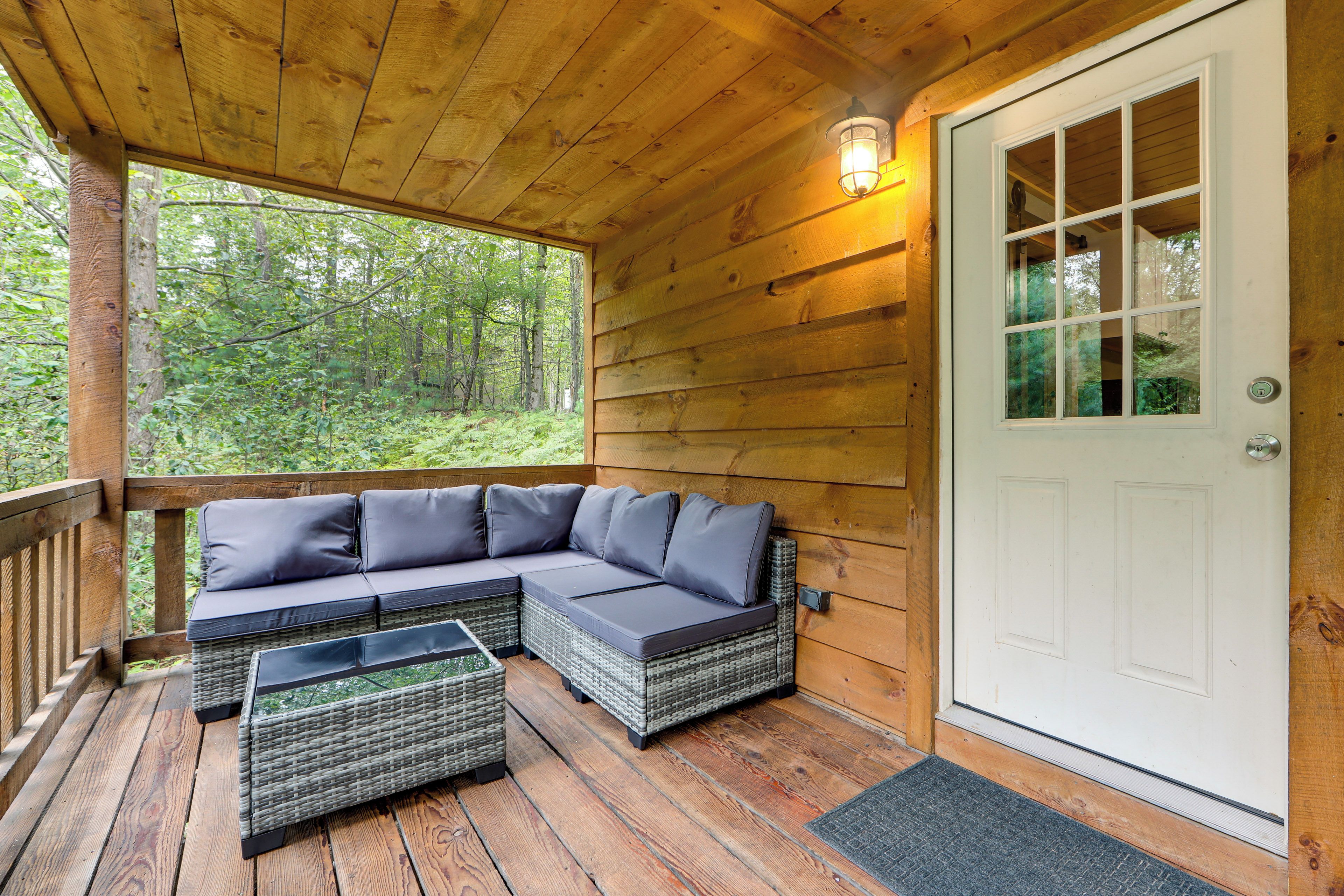 A wooden deck with a sectional sofa and wicker chairs, a door, and a view of a forest.