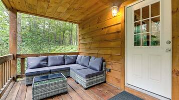 A wooden deck with a sectional sofa and wicker chairs, a door, and a view of a forest.