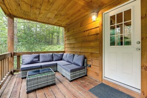 Une terrasse en bois avec un canapé modulable et des fauteuils en osier, une porte et une vue sur une forêt.