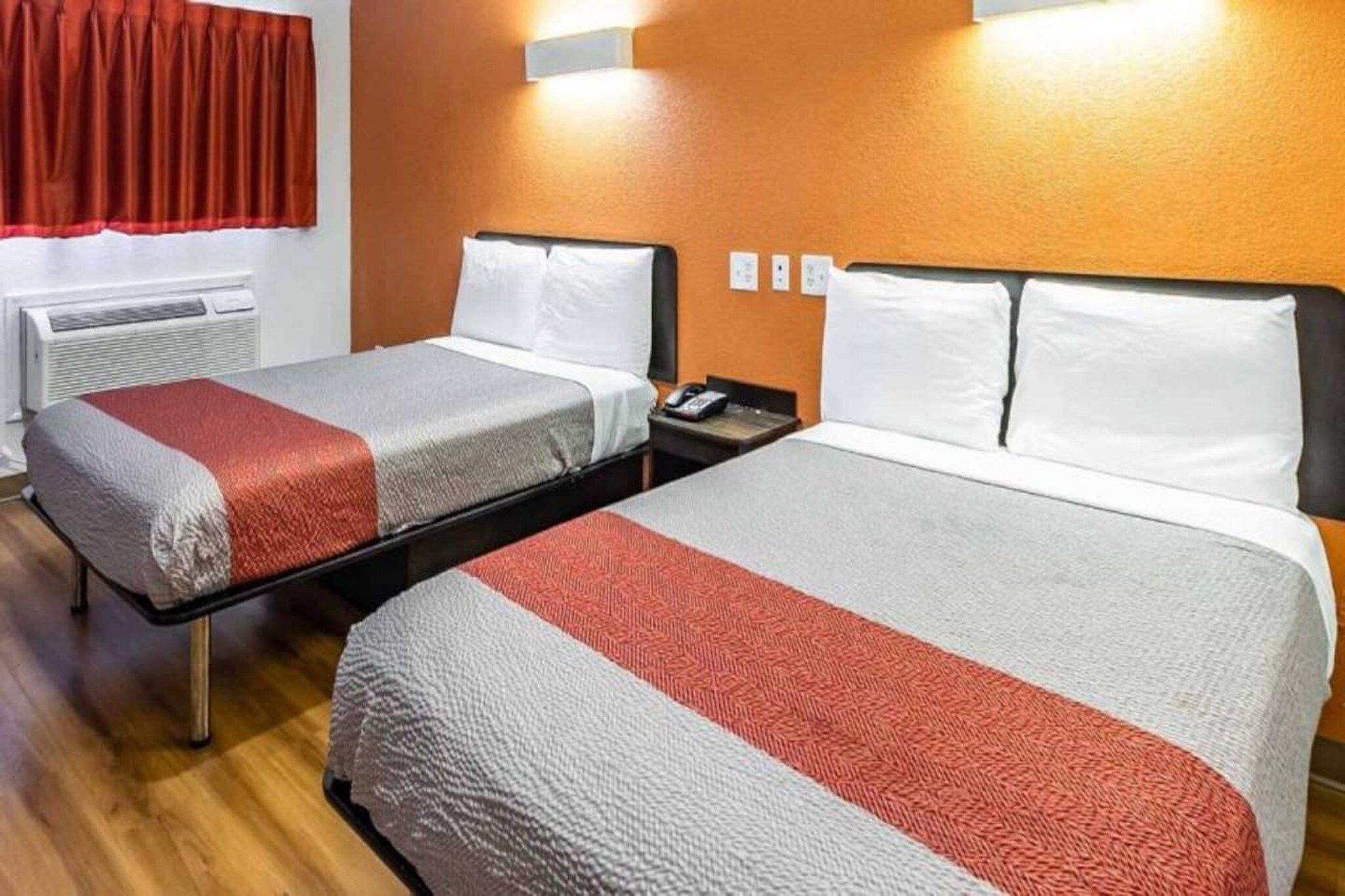 Habitación de hotel con dos camas, cada una con cubrecama blanco y rojo, una mesita de noche con teléfono y una lámpara fijada a la pared.