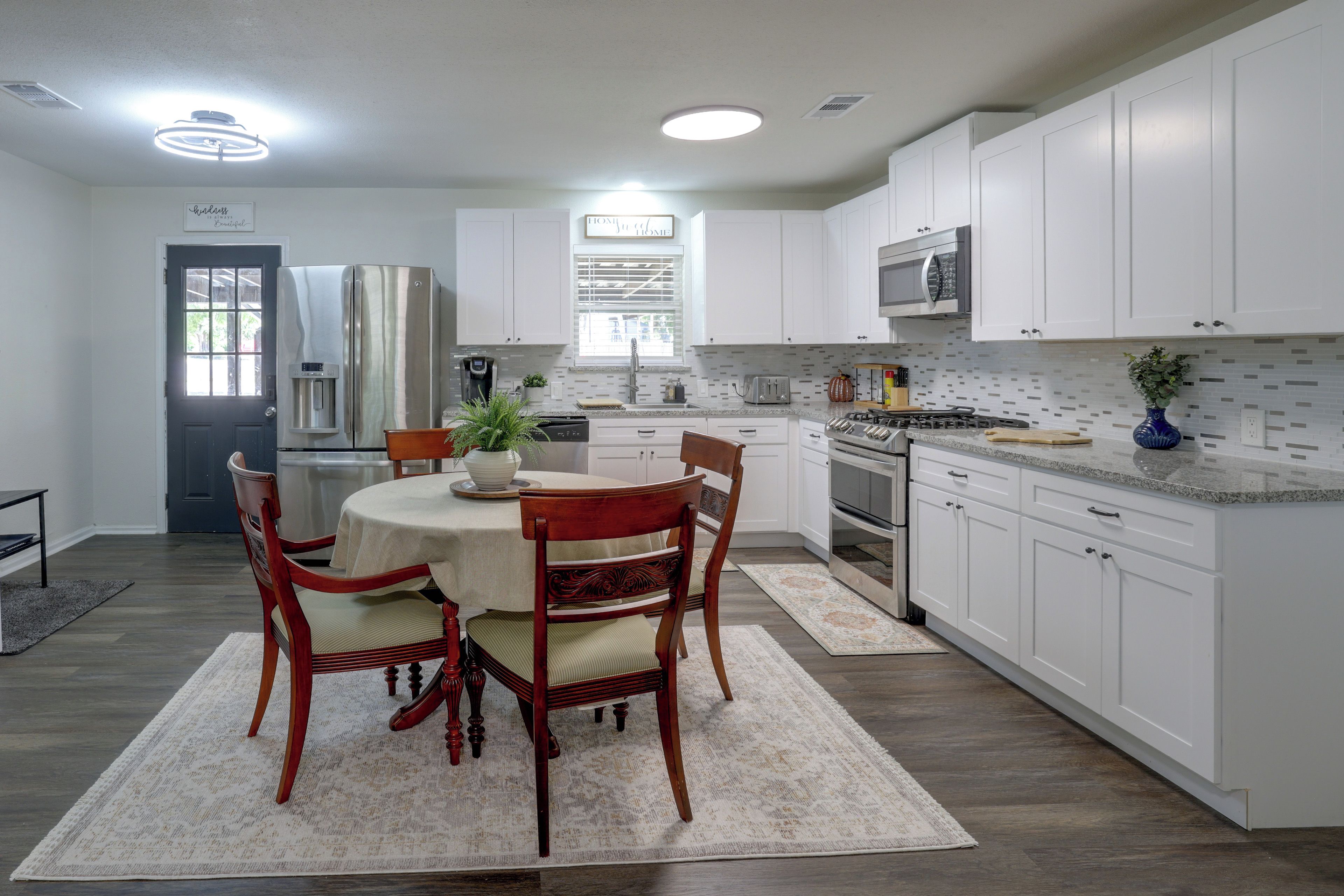 A modern kitchen with white cabinetry, a stainless steel refrigerator, a microwave, and a stove. There is a dining table with chairs and a rug.