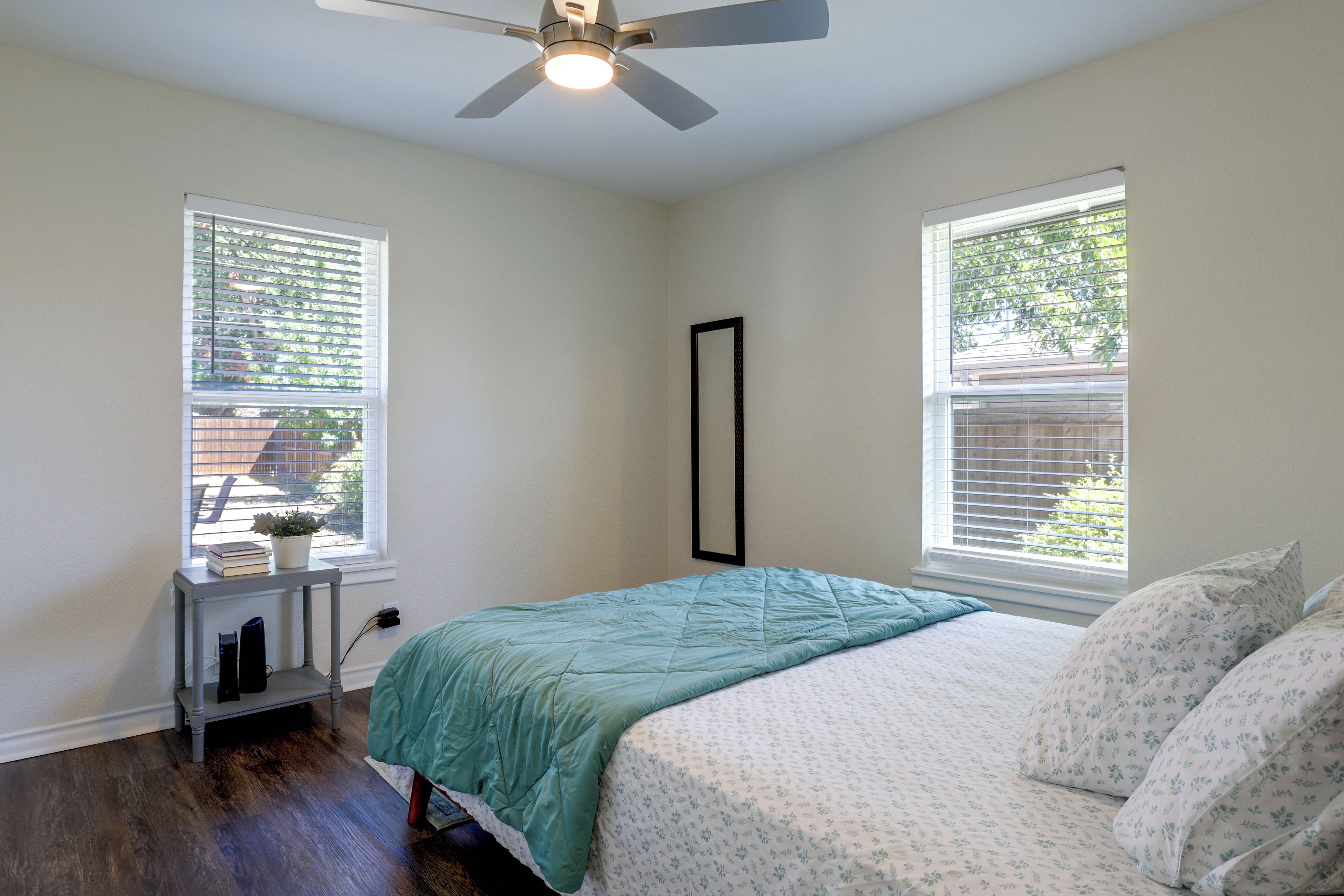 A bedroom with a bed, two windows, a ceiling fan, and a small table with a plant.