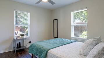 A bedroom with a bed, two windows, a ceiling fan, and a small table with a plant.