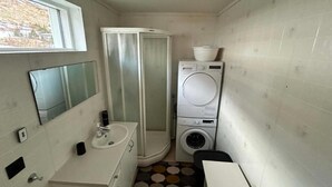 Ein kleines Badezimmer mit Waschbecken, Spiegel, Dusche sowie übereinander angeordneter Wasch- und Trockenmaschine.