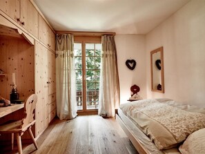 Ein Schlafzimmer mit einem Holzkleiderschrank, einem Schreibtisch mit Lampe, einem Bett mit gemustertem Überwurf und einem Fenster mit Vorhängen.